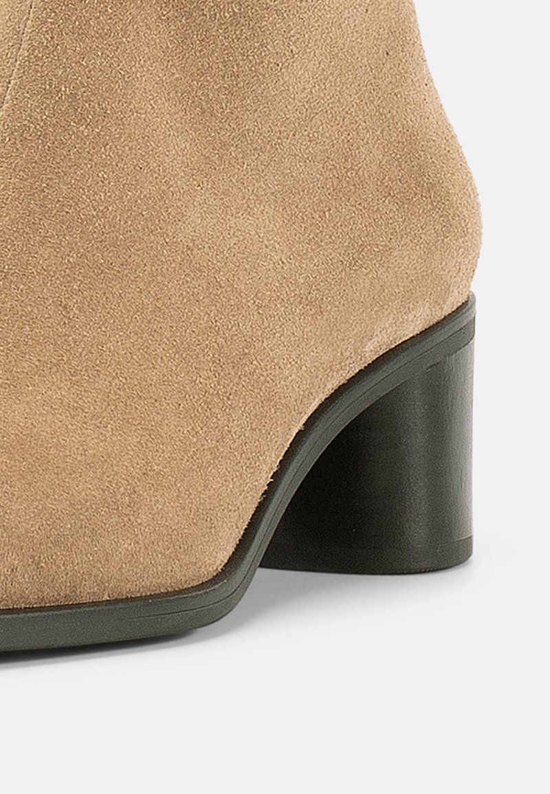 Beige ruskindsstøvle med rundet tå og en kraftig sort hæl. Glat tekstur uden synlige mønstre eller detaljer.