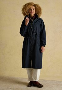 Palton lung impermeabilizat de culoare bleumarin, cu un cordon la talie, mâneci elastice și accente aurii. Purtați peste o cămașă în carouri și pantaloni bej.