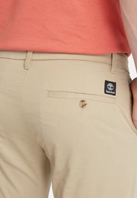 Pantaloni beige con una tasca posteriore dotata di bottone e etichetta con il logo Timberland. Il materiale ha una texture liscia.