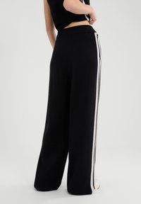 Schwarze, weit geschnittene Hose mit weicher Textur, weißen Seitenstreifen und hohem Bund, von hinten gezeigt.
