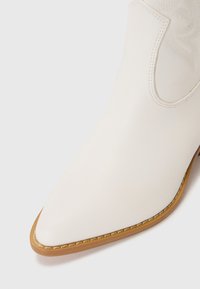 Bottines en cuir blanc avec une pointe effilée, semelle contrastante beige et couture décorative. Présente des découpes latérales et un motif texturé sur la tige.