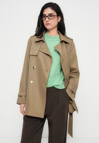 Καφέ χακί παλτό τύπου trench με χρυσά κουμπιά, μακριά μανίκια και ζώνη που δένει στη μέση. Φορεμένο πάνω από πράσινο πουλόβερ και καφέ παντελόνια. Ομαλή υφή υφάσματος.