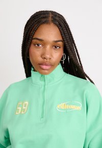 Felpa verde con mezzo zip, caratterizzata da un logo ricamato giallo e dal numero '59'. Tessuto testurizzato con lunghi capelli intrecciati in vista.