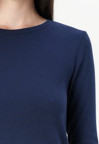 Nærbilde av en person som har på seg en ensfarget marineblå langermet genser med rund hals mot en hvit bakgrunn.