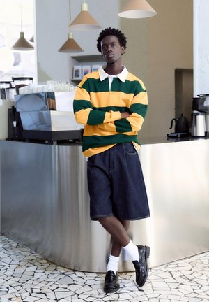 Mladić u žutozelenom prugastom džemperu i traper kratkim hlačama naslonjen na šank kafića s aparatima za kavu u blizini.