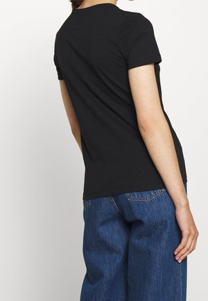 Person iført en ensfarvet sort t-shirt med korte ærmer og blå cowboybukser, vist bagfra mod en ensfarvet hvid baggrund.