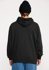 Sweatshirt noir à capuche avec une coupe décontractée, des poignets et un ourlet côtelés. Le matériau semble doux avec une texture lisse ; porté avec un jean bleu ample.