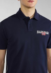 Tummansininen polo-paita, joka on valmistettu teksturoidusta kankaasta. Paidassa on kaksinappinen kaulus, lyhyet hihat ja rintaosassa lippumaisia kuvioita esittävä logo.