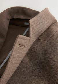 Gros plan d’un col de manteau en tissu brun texturé avec une boutonnière et un détail de couture structurée.