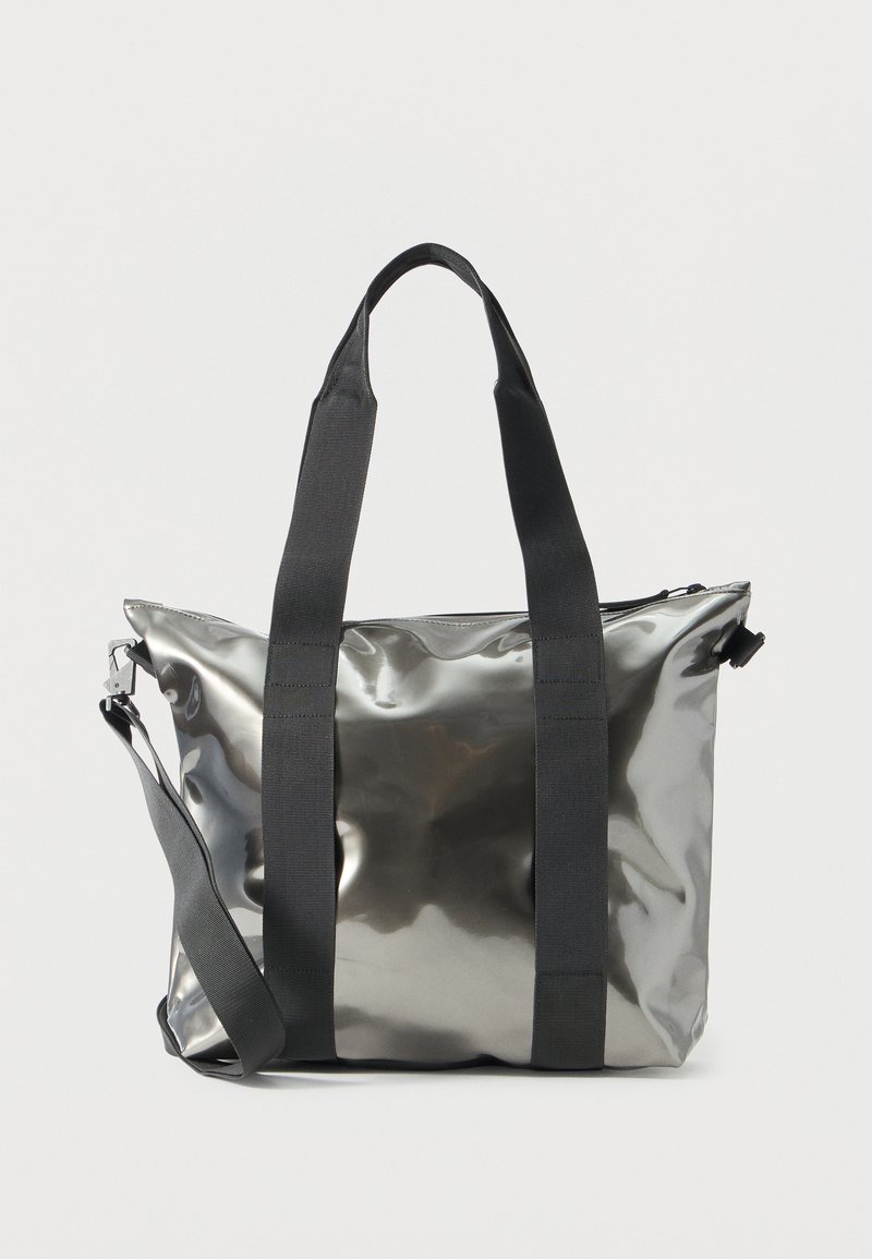 Λαμπερή μεταλλική ασημί τσάντα ώμου με φαρδιές μαύρες τιράντες και αποσπώμενο ιμάντα ώμου, εμφανίζεται πάνω σε απλό ανοιχτόχρωμο φόντο.