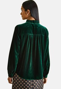 Grünes Samt-Hemd mit langen Ärmeln, das einen gerüschten Kragen, ein gerafftes Rückendesign und eine glatte Textur aufweist. Darunter ist ein dunkel gemusterter Rock sichtbar.