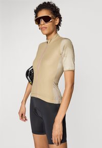 Beige sykkeltrøye med korte ermer og glidelås foran, med rosa detaljer; kombinert med svarte polstrede shorts og holder en svart hjelm.