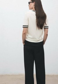 Woman with long brown hair wearing black sunglasses, a white short-sleeve top with black stripes, and black pants standing against a light wall.