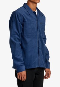 Camicia di velluto a coste blu navy con chiusura frontale a bottoni, due tasche sul petto e maniche lunghe. Presenta un colletto classico e una texture sottile.