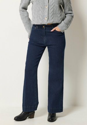Person iført mørkeblå jeans med høj talje og vidde ben, grå kabelstrikket knappet sweater, sorte ankelstøvler og hånd i lommen.