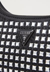 Svart tygväska med ett rutmönster av silver- och svarta rhinestones, en triangel av svart Guess-logotyp, samt ett dragkedjelås upptill.