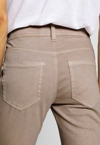 Primer plano de la parte trasera de una persona que lleva pantalones beige con dos bolsillos traseros y una camisa blanca metida por dentro.