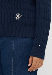 Ναυτικό πλεχτό πουλόβερ με ριγωτούς καρπούς, διαθέτοντας μια διακριτική κεντημένη λογότυπο και χρωματική λεπτομέρεια στο μανίκι. Συνδυάζεται με σκούρο τζιν.