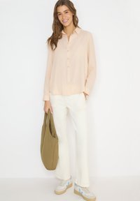 Femme portant un chemisier rose clair à boutons, un pantalon blanc et des baskets blanches, tenant un sac fourre-tout vert olive et souriante.