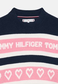 Mørk blå genser med rosa og hvite horisontale striper, med hjertemønstre og "TOMMY HILFIGER" tekst i fete bokstaver. Strikket tekstur.