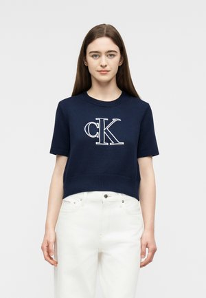 Młoda kobieta z długimi brązowymi włosami, ubrana w granatowy sweter z krótkim rękawem z białym logo "cK" oraz białe spodnie z wysokim stanem, stoi.