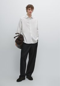 Witte overhemd met knopen, zwarte losse broek en donkere schoenen. Houdt een bruine suède tas vast. Effen achtergrond.
