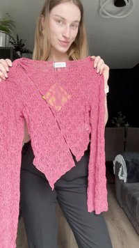 Top de encaje rosa de manga larga que presenta un patrón floral texturizado y un dobladillo desigual. Etiqueta visible en el interior del escote. Pantalones negros llevados por debajo.