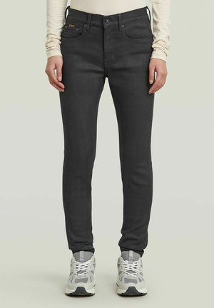 Persoon draagt slim-fit zwarte jeans en witte sneakers, staand tegen een effen lichte achtergrond met handen ontspannen langs het lichaam.