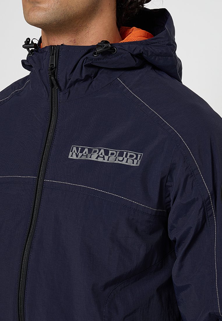 Giacca blu navy con chiusura a zip, cappuccio e fodera arancione a contrasto. Presenta un logo sul lato sinistro del petto e dettagli cuciti.