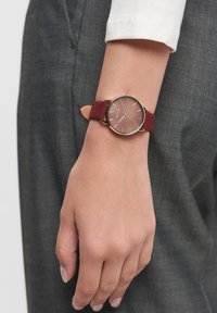Ronde horloge met een roségouden kast, kersenrode suède band en een lichtbruine wijzerplaat met gouden uurmarkeringen. Hand ligt op grijs textiel.