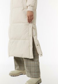 Gepolsterter cremefarbener Mantel mit seitlichen Knöpfen, kombiniert mit braunem Karomuster-Hosenanzug und beigefarbenen Stiefeln mit dickem Profil. Sanfte Textur und elegantes Design.