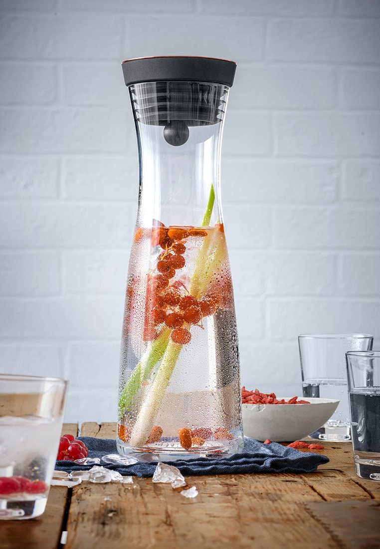 Carafe en verre transparent avec une forme conique, contenant du céleri vert et des baies rouges à l'intérieur, condensation sur la surface, et un couvercle noir.