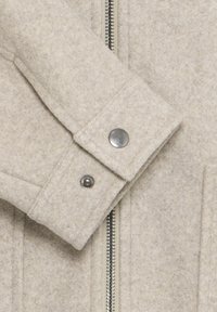 Beige uldjakke med en tekstureret overflade, der har en sølvfarvet lynlås, trykknaplukning og struktureret kravedetalje.