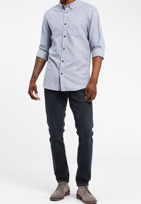 Mężczyzna w jasnoniebieskiej koszuli z guzikami i podwiniętymi rękawami, ciemnych jeansach slim-fit oraz szarych zamszowych butach na sznurówki, stojący na tle białej ściany.