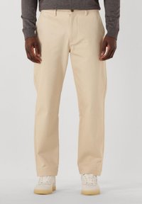 Bež krojene hlače ravnog kroja, izrađene od glatkog materijala, s prednjim zatvaračem i kopčanjem na dugme, uparenim s bijelim tenisicama.