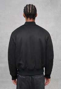 Giacca bomber in satin nero con collare e polsini a coste. Presenta un logo discreto sul retro e una texture liscia e lucida.