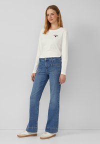 Weißes Langarmshirt mit kleinem schwarzem Grafikdruck, kombiniert mit hoch taillierten blauen Jeans im Schlag. Mit Front- und Gesäßtaschen akzentuiert.