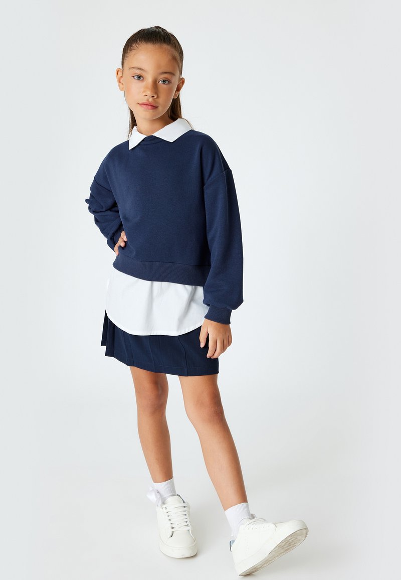 Navy Sweatshirt über einem weißen Hemd mit Kragen, kombiniert mit einem dunklen marineblauen gefältelten Rock und weißen Turnschuhen; entspanntes Design, weicher Stoff.