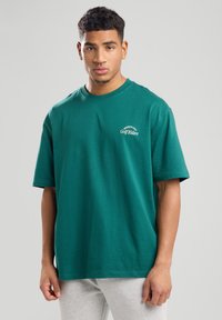 Vihreä puuvillateepaita, jossa on lyhyet hihat, pyöreä kaula-aukko ja valkoista brodeerattua tekstiä "Members Only Golf Riders" edessä.