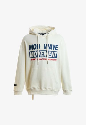Joven mirando al frente, vistiendo una sudadera crema con el texto "Mod Wave Movement", pantalones rojos y botas marrones sobre un fondo blanco liso.