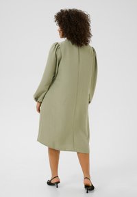 Robe midi vert olive avec des manches bouffantes, en tissu texturé, et une coupe droite. Présente un detailing sur la couture arrière. Des talons complètent la tenue.