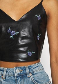 Czarny top z faux skórki o kroju crop z dekoltem w kształcie litery V. Zdobiony fioletowym haftem z kwiatami i zielonymi liśćmi. Połączony z jasnoniebieskimi dżinsami.
