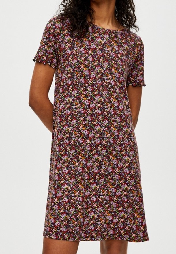 Femme portant une robe à manches courtes, de longueur genou, avec un motif floral dense en rouge, rose, violet et vert sur un fond sombre.
