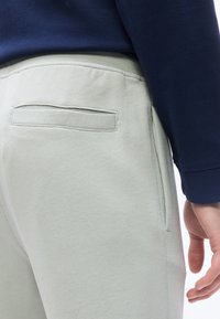 Κοντινό πλάνο τσέπης και πλαϊνής ραφής ανοιχτόγκρι παντελονιού, με ένα χέρι κοντά στον γοφό, φορεμένο με σκούρο μπλε μπλουζάκι με μακριά μανίκια.