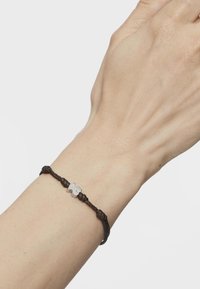 Pulsera de cordón trenzado marrón con un bead en forma de flor plateada en el centro, llevada en una mano sobre un fondo liso.