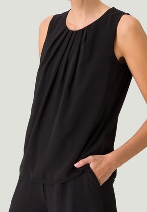 Haut noir sans manches avec des plis au niveau de l'encolure, porté par une personne ayant une main dans la poche d'un pantalon noir assorti.