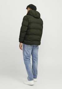 Chaqueta acolchada verde oscuro con capucha; diseño acolchado, puños elásticos y corte relajado, llevada sobre unos vaqueros azul claros y zapatillas blancas.