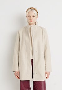 Beige ullblandning kappa med hög krage, dragkedja och tryckknappar. Har rak skärning och långa ärmar, kombinerad med en beige tröja och vinröda byxor.