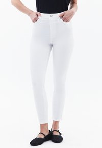 Jeans blancos de cintura alta con corte ajustado, que cuentan con bolsillos delanteros y cierre de botón metálico. Combinados con zapatos tipo slip-on de malla negra.