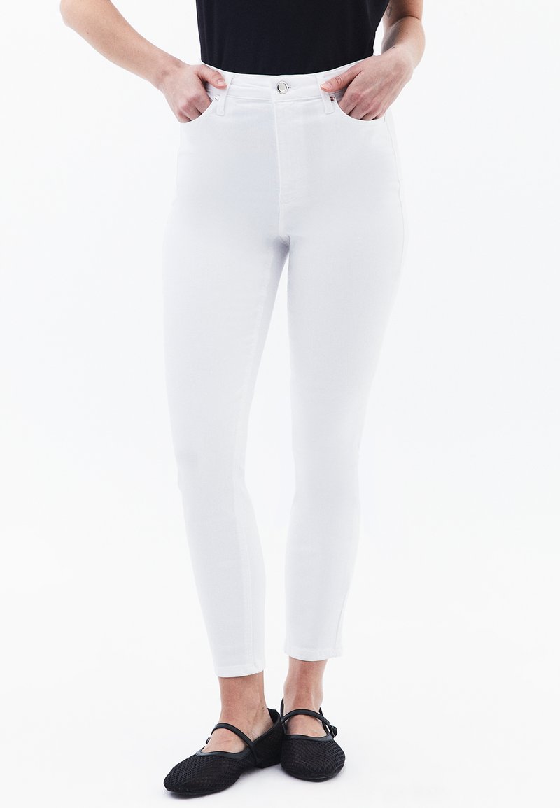 Jeans blancos de cintura alta con corte ajustado, que cuentan con bolsillos delanteros y cierre de botón metálico. Combinados con zapatos tipo slip-on de malla negra.